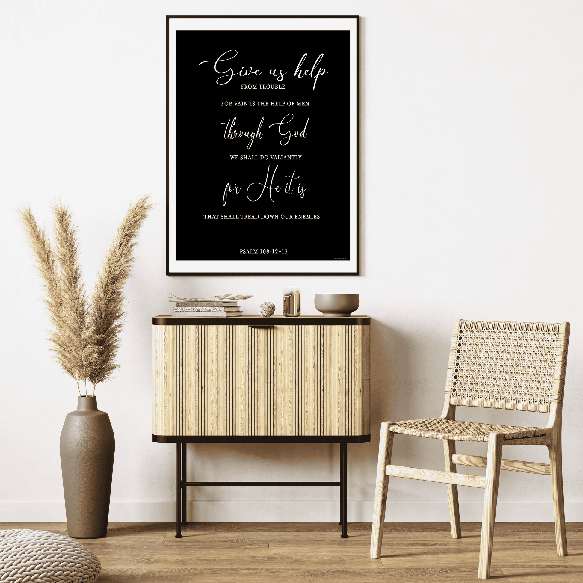White-painted room featuring a wicker chair, a wood and rattan cabinet adorned with decorative items on top, dried plants in a vase, and the Psalm 108:12-13 Art Print by Olive Grove Life framed on the wall.