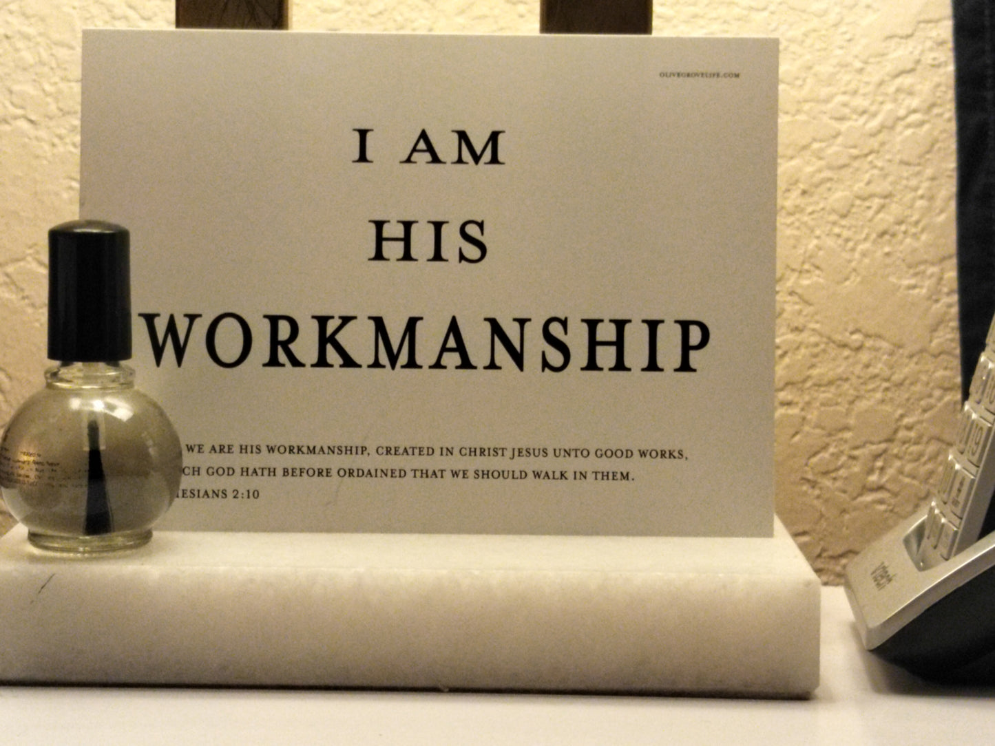 Card with text 'I AM HIS WORKMANSHIP' on a surface with a bottle and phone.