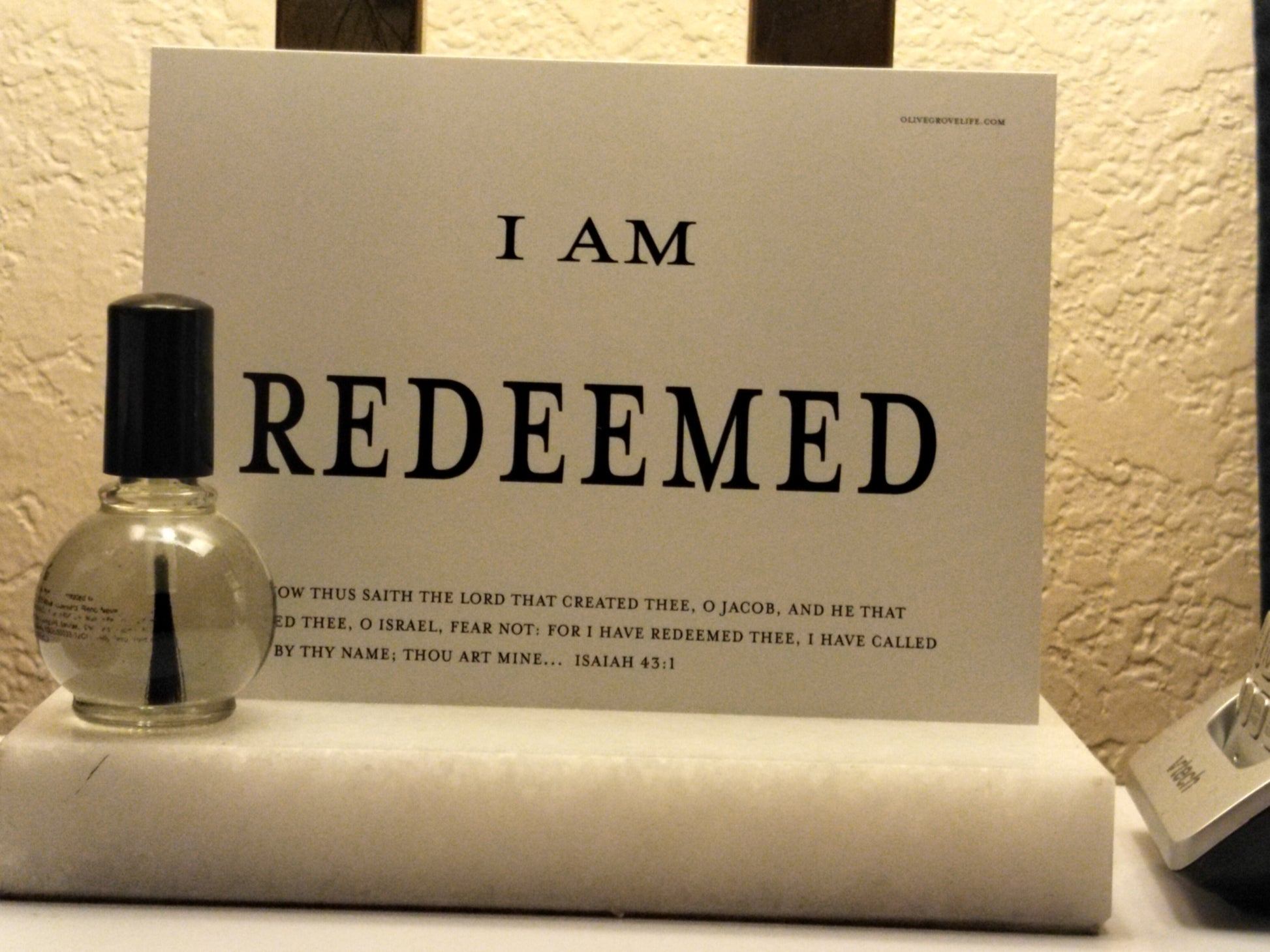 Small bottle of perfume next to a card with 'I AM REDEEMED' text on a textured surface.
