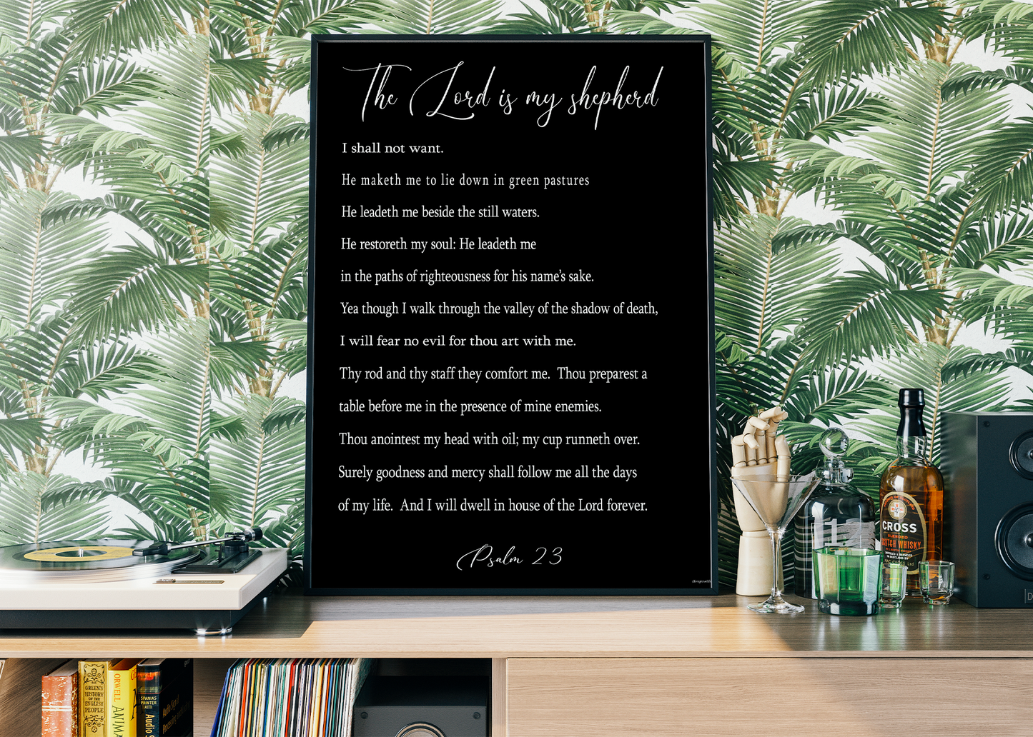 The Psalm 23 Art Print by Olive Grove Life is framed and set on a wooden shelf against vibrant tropical wallpaper.